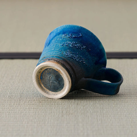 Blue glazed mug tipped over to show its foot and handle, on a tatami mat.
