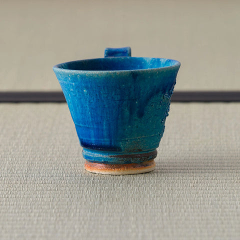 Tall deep blue glazed cup with a small lug handle, viewed showing the interior, on a tatami mat.