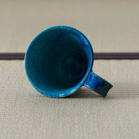 Blue glazed mug tipped over to show its swirled blue interior and handle, on a tatami mat.