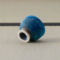 Deep blue glazed cup tipped over to show its foot, on a tatami mat.