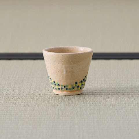 Sandy stoneware cup with a green-and-blue dotted band, side view on a tatami mat.