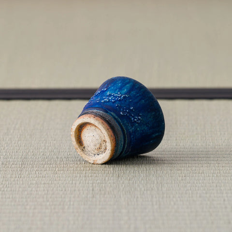 Deep blue speckled cup tipped over to show its pale foot, on a tatami mat.