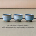 Three pale blue teacups in a row with a note that handmade pieces vary slightly, on a tatami mat.