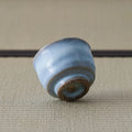 Pale blue crackle-glazed ceramic teacup tipped over to reveal its unglazed foot, on a tatami mat.