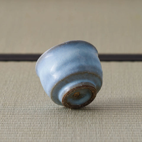 Pale blue crackle-glazed ceramic teacup tipped over to reveal its unglazed foot, on a tatami mat.