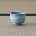 Pale blue crackle-glazed ceramic teacup upright on a tatami mat, three-quarter view.
