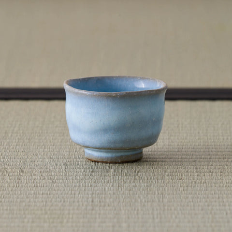 Pale blue crackle-glazed ceramic teacup upright on a tatami mat, three-quarter view.