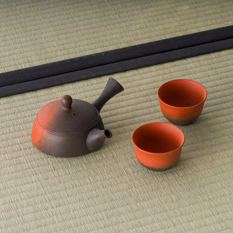 Two-tone tokoname tea set — a side-handle kyusu with a dark top and reddish-clay base, shown with two matching red cups on tatami