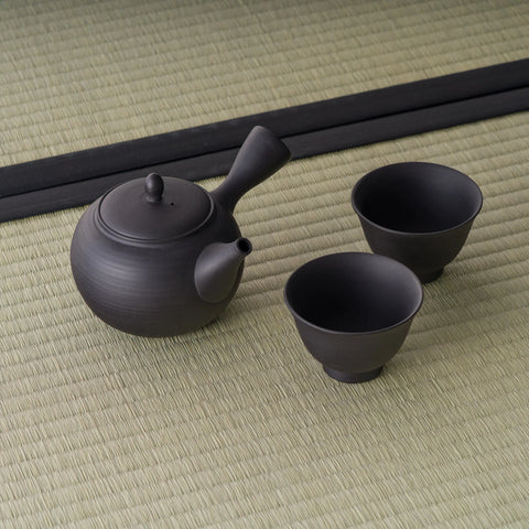 Black unglazed tokoname-style tea set — a side-handle kyusu teapot with two matching cups, arranged on tatami