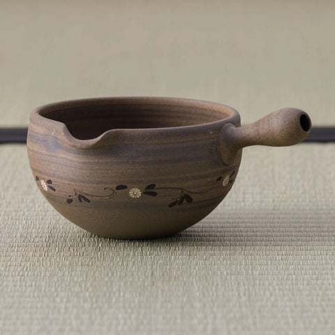 Brown stoneware spouted bowl (katakuchi) with an inlaid floral band and a wooden handle, side view on a tatami mat.