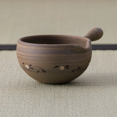 Brown stoneware spouted bowl (katakuchi) with an inlaid floral band and a wooden handle, on a tatami mat.