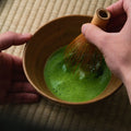 A hand whisking bright green matcha with a bamboo chasen in a brown ceramic bowl, on a tatami mat.