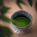 Prepared green matcha in a rustic speckled tea bowl on a wooden table, with leaf shadows falling across it.