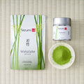 Tezumi Wakatake matcha pouch printed with green bamboo beside a silver tin and a small dish of matcha powder on a tatami mat.