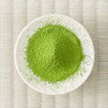 Green Wakatake matcha powder mounded in a white dish on a tatami mat, overhead view.