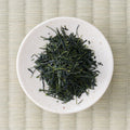 Long slender Wakō sencha leaves on a small white dish, seen from directly above on tatami