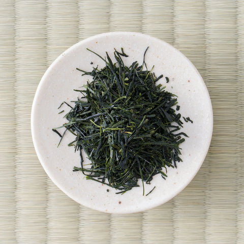 Long slender Wakō sencha leaves on a small white dish, seen from directly above on tatami