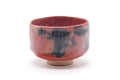 Red aka-raku chawan with dark grey-blue patches and a pale foot, shown from a three-quarter angle.