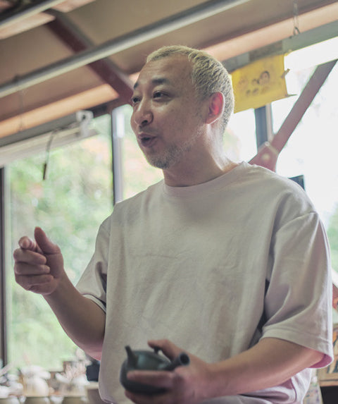 Potter Tsuzuki Yutaka standing in his workshop, gesturing as he speaks, with tools and shelves behind him