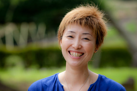Shiho Sato smiling outdoors in a blue blouse, with blurred green foliage behind her.