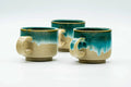 Set of three cups with a turquoise drip glaze fading to pale beige clay, grouped together.