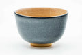 Side profile of the grey celadon chawan showing a decorative floral-and-kanji roundel and the beige glazed interior.
