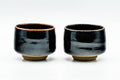 Pair of small jet-black glazed yunomi side by side, front view with a lighter metallic break at the rim and exposed buff foot.