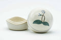 Round white kogo (incense container) with its lid propped beside the base, hand-painted with a blue flower and green leaves.