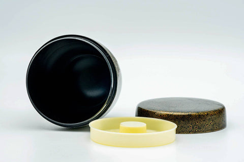 The canister open showing its dark interior, with the cream inner lid and speckled outer lid lying flat beside it.