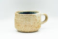 Beige speckled ushirode handled yunomi at a three-quarter angle, small loop handle on the right and a cerulean-blue glazed interior visible at the rim.