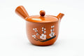 Three-quarter front view of a red shudei Tokoname-yaki kyusu decorated with engraved white ume plum blossoms, lid on, side handle right and spout left.