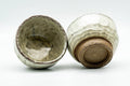 A pair of milky-beige wabi-sabi yunomi, one upright and one tipped on its side to reveal the unglazed clay foot.