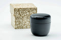 Small black-urushi matcha tea caddy (ko-natsume) shown beside its speckled stone-textured storage box, lid on