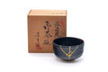 Black matcha bowl (chawan) with gold kintsugi repair, shown beside its signed wooden storage box.