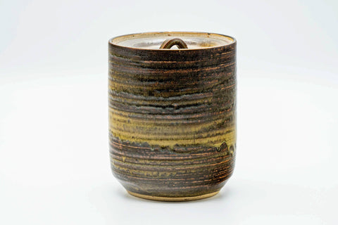 Front view of the lidded brown-beige mizusashi rotated slightly, highlighting the streaked horizontal glaze around the cylindrical body.