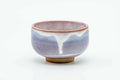 Small Hagi-yaki teacup with a lavender-purple glaze and white drips, raised on a beige foot, front view.