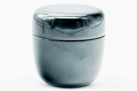 Glossy black-urushi matcha tea caddy, lid on, three-quarter close view with a light highlight on the black lacquer