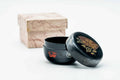 Black-lacquer matcha tea caddy (natsume) with gold floral maki-e and a red crest, shown beside its wooden storage box, lid on