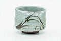 Front view of a cylindrical matcha bowl in pale grey-green glaze with dark grass-like drip brushwork down one side and an exposed clay foot.