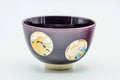 Front view of a deep aubergine-purple Kyo-yaki matcha bowl decorated with round medallions of gold-and-blue geometric patterns on a pale foot.