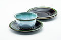 Stack of black lacquer saucers with green-trimmed rims and a celadon-glazed teacup on top for scale, three-quarter view.