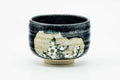 Small black metallic-glazed matcha chawan with a painted white floral spray on a beige panel, three-quarter view