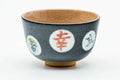 Grey celadon matcha bowl (chawan), three-quarter view, exterior decorated with white roundels containing red kanji and coloured florals, warm beige glazed interior rim visible.